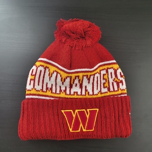 May include: A red knit beanie with a matching pom-pom. The word "COMMANDERS" is across the crown in white and gold lettering. The team's "W" logo is embroidered in gold on the front.