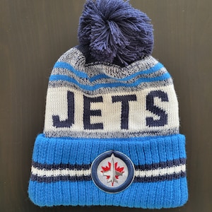 May include: A blue and white knit beanie with a pom pom on top. The beanie has the word "JETS" in black letters on the front and a blue and white logo with a maple leaf on the side.