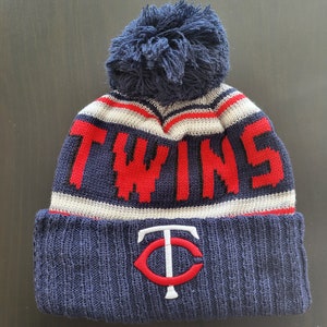 May include: A navy blue knit beanie with a large pom-pom. The word "TWINS" is knit in red across the crown. The front of the hat features a red and white logo. A winter hat for sports fans.