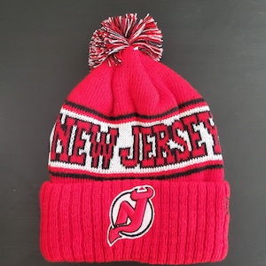 May include: Red knit beanie with the words "NEW JERSEY" in white block letters. The beanie features a pom-pom and a logo of a red "J" inside a white and black outline. A winter hat.