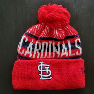 May include: A red knit beanie with the word "CARDINALS" in white letters across the top. The lower portion of the hat is red with the St. Louis Cardinals logo embroidered in white. A red pom-pom sits atop the hat.