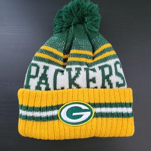 May include: A knitted winter hat in green, white, and yellow, featuring the word "PACKERS" in white block letters. The hat has a green pom-pom on top and a green and white logo patch with a "G" on the front. The brim is yellow with green and white stripes.