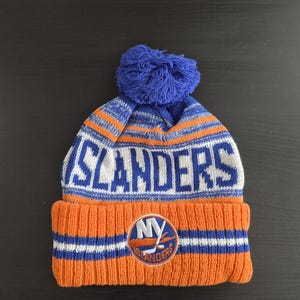 May include: An orange, blue, and white knit beanie with the word "ISLANDERS" in white block letters. The hat features a blue pom-pom and a patch with "NY ISLANDERS" on the front. A winter hat for sports fans.