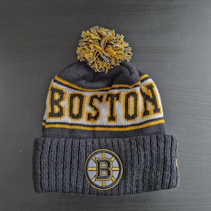 May include: A dark gray knit beanie with the word "BOSTON" in yellow and white block letters. The hat features a yellow and black pom-pom and a circular logo with a "B" in the center.