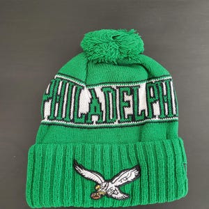 May include: Green knit beanie with the word "PHILADELPHIA" in white and black lettering. Features a green pom-pom and an embroidered eagle logo. A winter hat for fans of the Philadelphia Eagles.