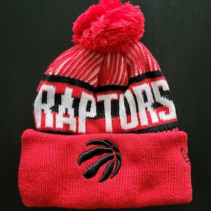 May include: Red and black knit beanie with the word "RAPTORS" in white block letters. The beanie features a red pom-pom and a black embroidered basketball logo. A New Era tag is visible.