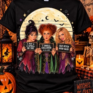 May include: A black short-sleeve t-shirt featuring a graphic of three women in witch costumes standing before a full moon. They hold signs that read I PUT A SPELL ON YOU, I SMELL CHILDREN 10-31-1693, and BAD WITCH GOOD HAIR, with bats and stars.