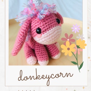 May include: A pink crocheted donkeycorn with a blue and pink mane and a white horn. The donkeycorn is sitting on a wooden surface with yellow and pink flowers.