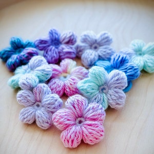 May include: A collection of crocheted flower embellishments in various shades of blue, purple, pink, and white. The flowers are arranged in a triangular shape on a light wood surface.
