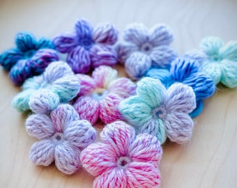 May include: A collection of crocheted flower embellishments in various shades of blue, purple, pink, and white. The flowers are arranged in a triangular shape on a light wood surface.