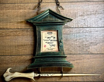 Puede incluir: Un marco de madera verde con una cita en su interior que dice "No "mora" en sueños y olvida "vivir"." El marco está colgado en una pared de madera con una varita colgando debajo.