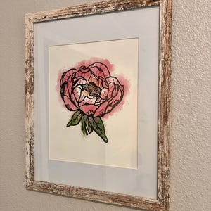 Puede incluir: Pintura de acuarela enmarcada de una flor de peonía rosa con hojas verdes. La obra de arte está sobre un fondo claro y enmarcada en un marco de madera clara envejecida. La pintura está montada sobre un passe-partout blanco.