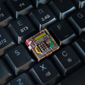 Könnte beinhalten: Nahaufnahme einer kleinen, detaillierten Leiterplatte, die auf einer schwarzen Computertastatur liegt. Die Leiterplatte weist verschiedene farbige Komponenten auf, darunter Grün, Gelb und Rot, und ist von schwarzen Tasten mit weißer Beschriftung umgeben.