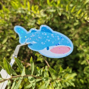 Può includere: Un adesivo a forma di balena a cartoni animati blu e rosa con dettagli glitterati. L'adesivo è tenuto da una clip in metallo argentato, su uno sfondo di fogliame verde. La balena ha un corpo azzurro con pinne blu scuro e una bocca rosa.