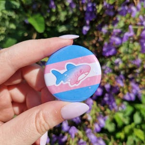 Peut inclure: Un bouton rond avec un motif bleu, rose et blanc, représentant un requin de dessin animé. Le fond du bouton reprend les couleurs du drapeau transgenre. Le bouton est tenu devant un fond de fleurs violettes.