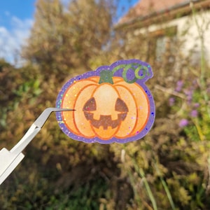 Peut inclure: Autocollant citrouille orange pailleté avec un visage souriant, une tige verte et une bordure violette. L'autocollant est tenu par une pince à épiler sur un arrière-plan extérieur flou, suggérant le thème d'Halloween.