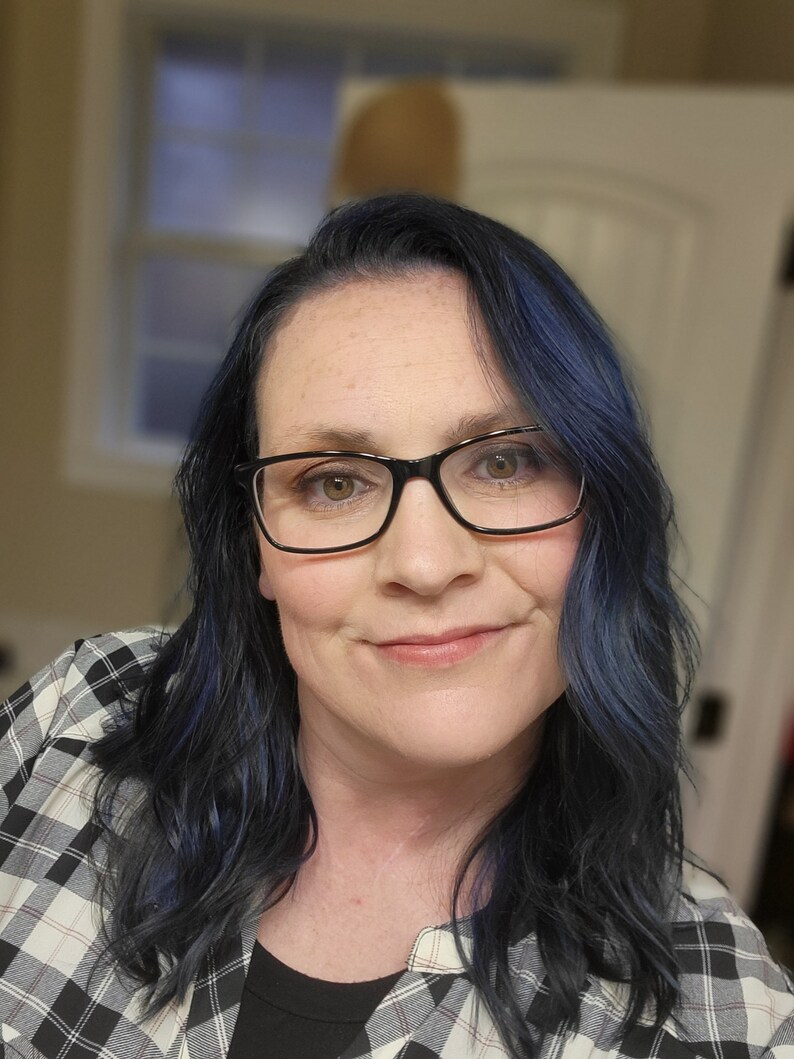 May include: A woman with dark blue hair wearing black-framed glasses and a black and white plaid shirt.