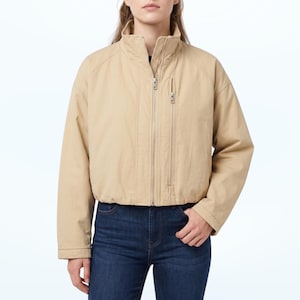 May include: A beige cropped jacket featuring a high collar, a full-length front zipper, and a vertical zippered chest pocket. The jacket has long sleeves with a relaxed fit and is paired with dark blue denim jeans, creating a neutral ensemble.