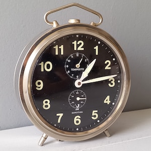 Peut inclure: Un réveil vintage avec un cadran noir et un boîtier en métal argenté. L'horloge présente de grands chiffres et aiguilles lumineux, avec un cadran plus petit en dessous. Le mot "TEMPESTO" est visible sur le cadran.