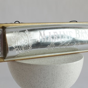 May include: A silver harmonica with the words "Marine Band" and "M. Hohner" engraved on the top. The harmonica is sitting on a white, textured surface.