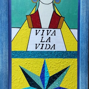Puede incluir: Una pintura enmarcada con fondo amarillo y una estrella azul y verde en el centro. La pintura presenta a una mujer con cabello negro y labios rojos. El texto "Viva La Vida" está escrito en negro sobre un fondo blanco.