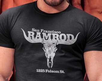 Pode incluir: Camiseta preta com um gráfico branco de um crânio de touro e o texto "San Francisco RAMROD" acima e "1225 Folsom St." abaixo. A camiseta é de manga curta.