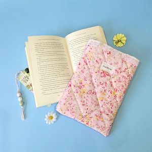 May include: A quilted fabric book sleeve featuring a pink floral pattern with yellow and green accents, a white lace trim, and a small rectangular label reading "LucyLocket". An open book and a beaded bookmark with a tassel are placed nearby.