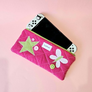 May include: A quilted hot pink fabric pouch with white polka dots, featuring a light green star appliqué, a white butterfly appliqué, and two pink buttons. A white gaming console with black screen and character decals is tucked inside the pouch.