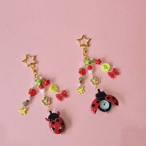 May include: Two gold-toned star-shaped keychains featuring dangling charms including red ladybirds, one with a functional clock face, green and red beads, small white pearls, yellow butterfly charms, lime green buttons, and red bows.