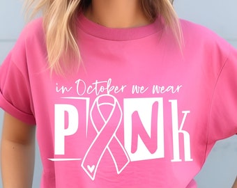 May include: A pink t-shirt with the text "in October we wear PINK" in white. The design includes a pink ribbon and a small heart. The shirt has rolled-up sleeves.