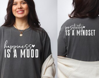 May include: A dark gray t-shirt with the text "happiness is a mood" on the front and "positivity is a mindset" on the back. Both phrases are in white text with a small heart symbol.