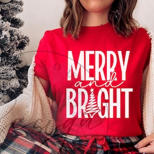 以下が含まれることがあります： 「MERRY and BRIGHT」の白い文字とクリスマスツリーのデザインが施された赤いTシャツ。赤と黒のチェック柄のパジャマパンツとクリーム色のセーターを着ています。背景には装飾されたクリスマスツリーがあります。