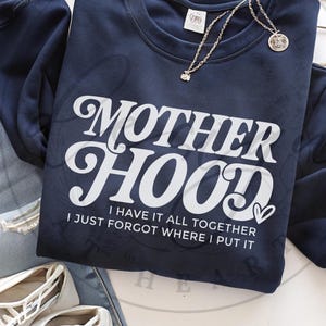 Op de afbeelding: Een marineblauw sweatshirt met witte tekst die "MOTHERHOOD I HAVE IT ALL TOGETHER I JUST FORGOT WHERE I PUT IT" luidt. De tekst is in een retro-lettertype met een hartje aan het einde van de zin.
