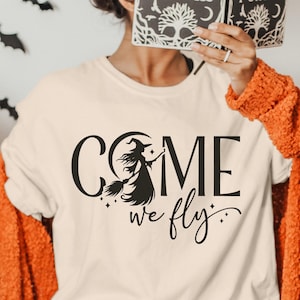 Peut inclure: T-shirt crème avec l'inscription noire "COME we fly". Le design intègre une silhouette de sorcière sur un balai, un croissant de lune et des étoiles. Ce t-shirt fait partie d'une collection sur le thème d'Halloween.