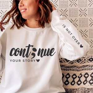 May include: White long-sleeved sweatshirt with the text "continue your story" and a semicolon symbol. The right sleeve has the text "is not over" with a small heart symbol.