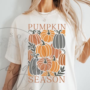 May include: Cream-colored t-shirt featuring a rectangular design with the words "PUMPKIN SEASON" in brown. The design includes various orange and gray pumpkins with green leaves, creating a fall-themed graphic.