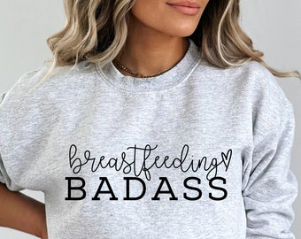 May include: A gray sweatshirt with the text "breastfeeding BADASS" in black lettering. The text is in a cursive font with a small heart symbol after the word "breastfeeding".