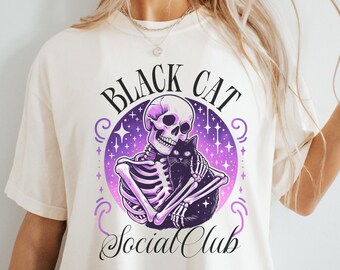 May include: Cream-colored t-shirt featuring a graphic of a skeleton embracing a black cat. The design is set within a purple and white circular frame with the words "BLACK CAT Social Club". The design has a spooky, gothic theme.
