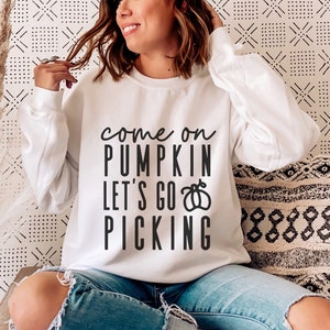 May include: White sweatshirt with the text "come on PUMPKIN let's go PICKING" in black. The sweatshirt is a crew neck style. The person in the image is wearing blue jeans.