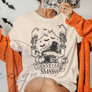 May include: Cream-colored t-shirt featuring a black graphic with skeletons, bats, pumpkins, and a vintage gramophone. The text reads "IT WAS A GRAVEYARD SMASH" in a banner. The shirt is part of a Halloween-themed collection.