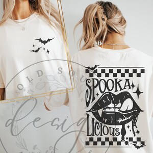 May include: Cream-colored t-shirt featuring a black bat and star design on the front. The back of the shirt displays the words "Spooka Licious" with a vampire mouth graphic, bats, and a checkered pattern, perfect for Halloween.