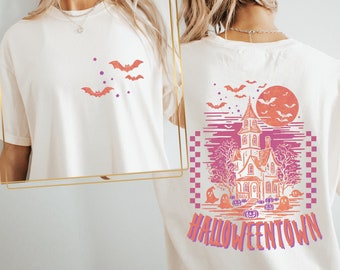May include: Cream-colored t-shirt featuring a Halloween design. The front displays orange bats and purple stars. The back showcases a haunted house, full moon, bats, ghosts, and the word "Halloweentown" in orange and purple.