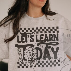 May include: A light gray sweatshirt with the words "LET'S LEARN TODAY" in a black, retro font. The design includes a pencil and a smiling flower. The top has a checkered pattern.