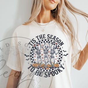 May include: Cream-colored t-shirt with a Halloween-themed graphic. The design features three dancing skeletons, pumpkins, and the text "'Tis the Season to be Spooky" in a circular checkered frame. Bats are also included in the design.
