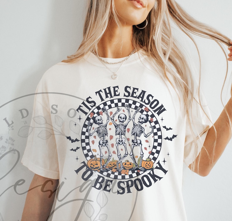 May include: Cream-colored t-shirt with a Halloween-themed graphic. The design features three dancing skeletons, pumpkins, and the text "'Tis the Season to be Spooky" in a circular checkered frame. Bats are also included in the design.