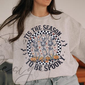 May include: A light gray sweatshirt with a Halloween-themed graphic. The design features three dancing skeletons inside a checkered circle, surrounded by pumpkins and bats. The text reads "TIS THE SEASON TO BE SPOOKY."