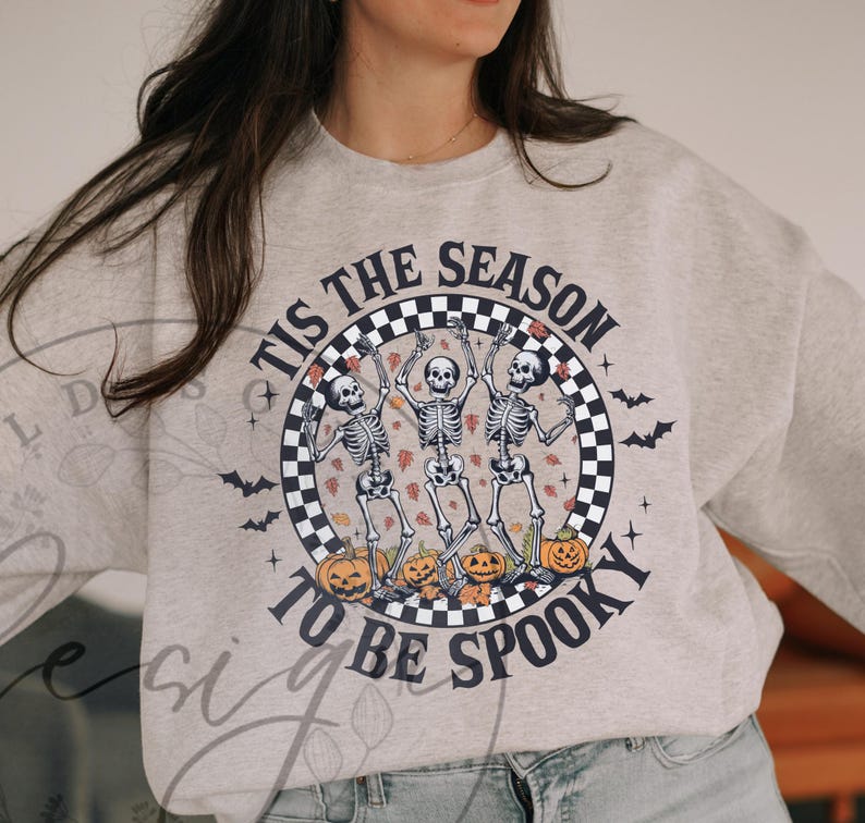 May include: A light gray sweatshirt with a Halloween-themed graphic. The design features three dancing skeletons inside a checkered circle, surrounded by pumpkins and bats. The text reads "TIS THE SEASON TO BE SPOOKY."