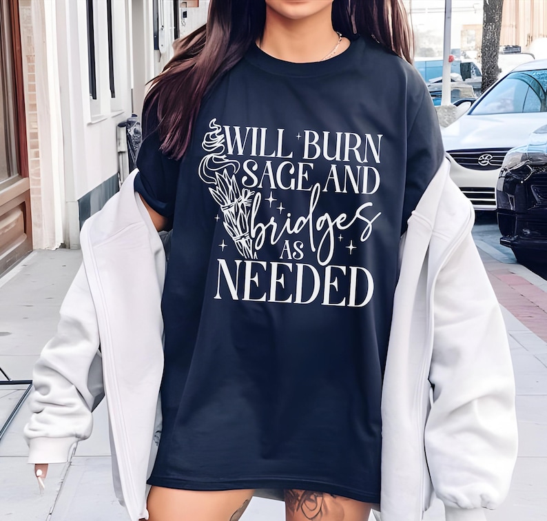 May include: A dark blue t-shirt with white text that reads "Will burn sage and bridges as needed". The text is surrounded by a stylized image of a burning sage bundle.