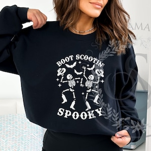 May include: Black sweatshirt with a white graphic that reads "Boot Scootin' Spooky." The design features dancing skeletons, bats, and stars, perfect for Halloween. A crew neck sweatshirt.