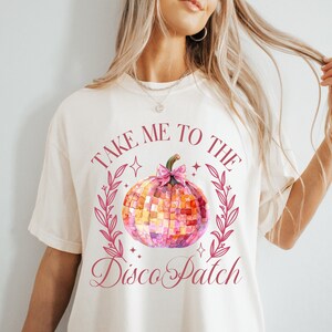 May include: Cream-colored t-shirt featuring the text "TAKE ME TO THE Disco Patch" in a burgundy script. A watercolor disco ball with pink and orange squares is centered, surrounded by a burgundy wreath. The shirt has a relaxed fit.